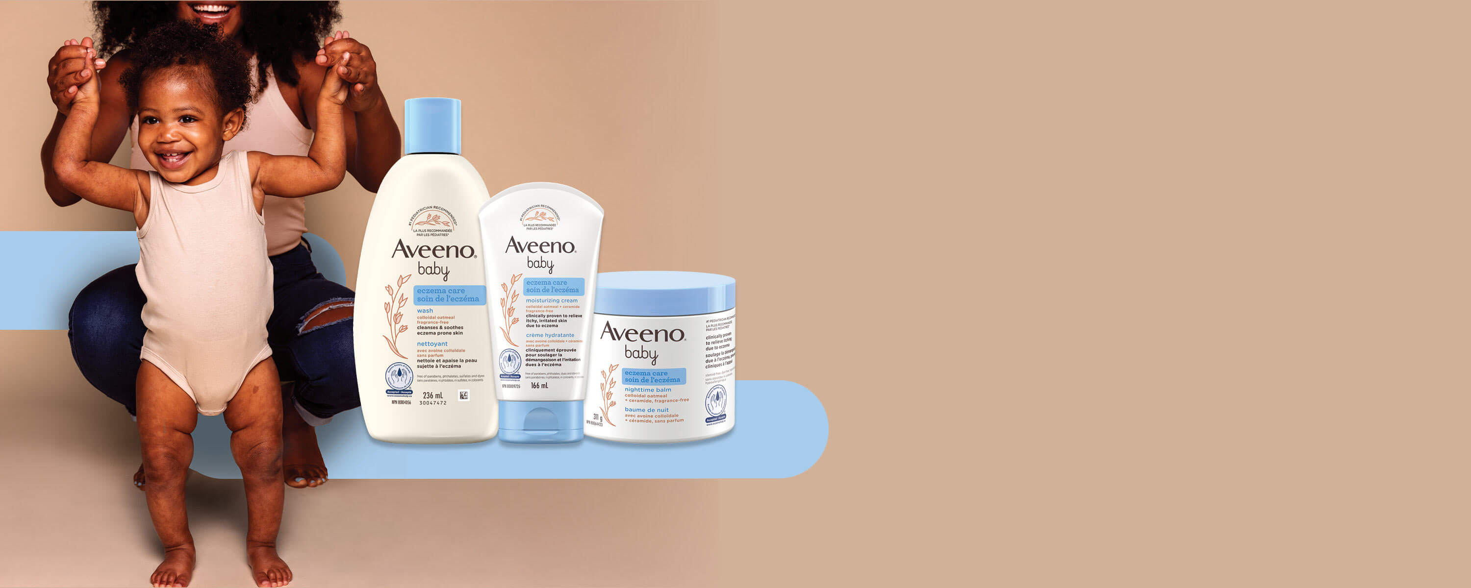 Young mother holding her toddler’s hand while they are standing and smiling with 3 Aveeno Baby eczema products next to them.