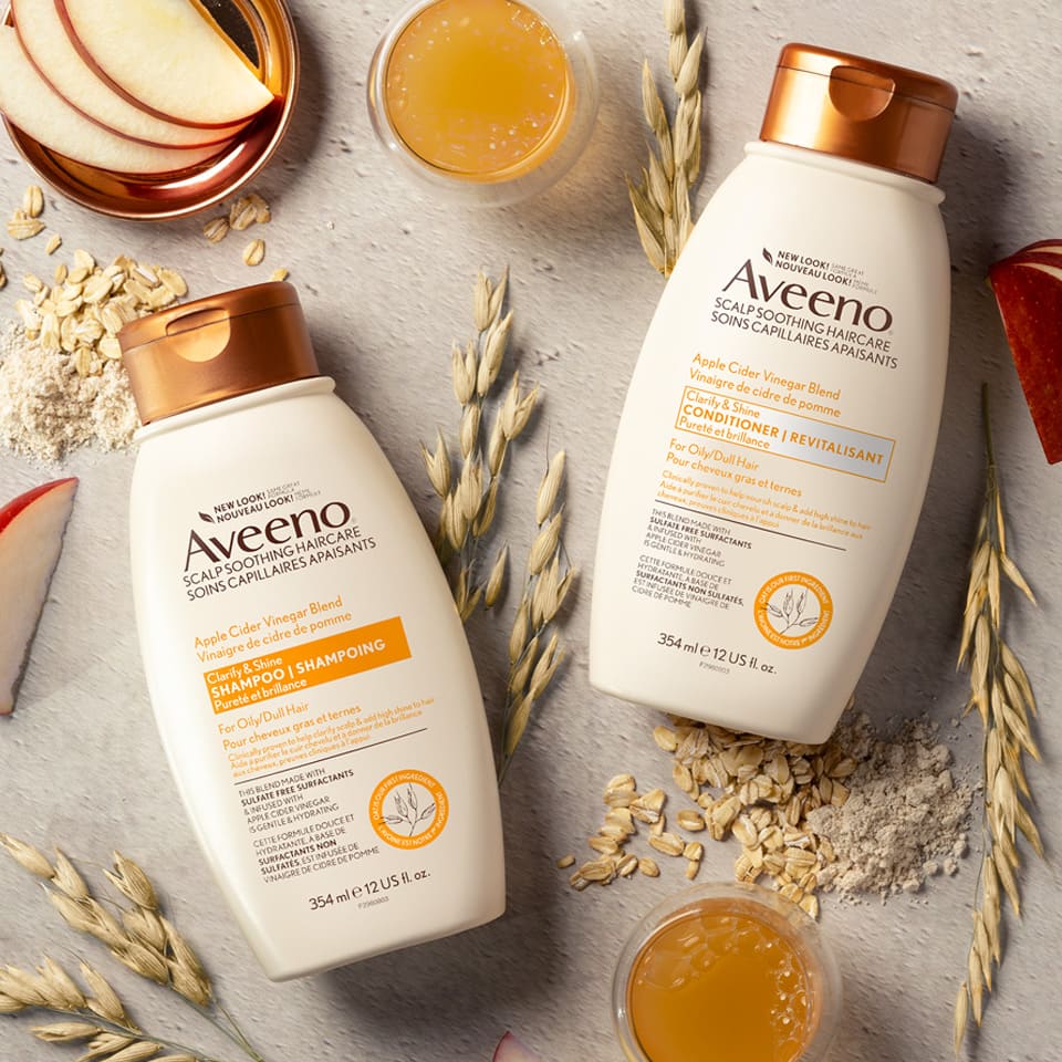 AVEENO® Apple Cider Vinegar Blend Clarify and Shine Hair Conditioner and Shampoo bottles with oat, apple slices and cup of vinegar in the background