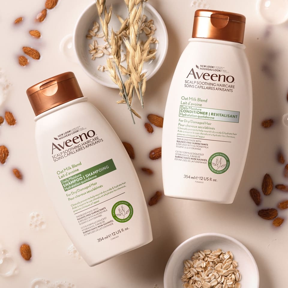 AVEENO® Oat Milk Blend Daily Moisture Hair Shampoo and Conditioner Bottles with oat and almonds soaked in oak milk in the background.
