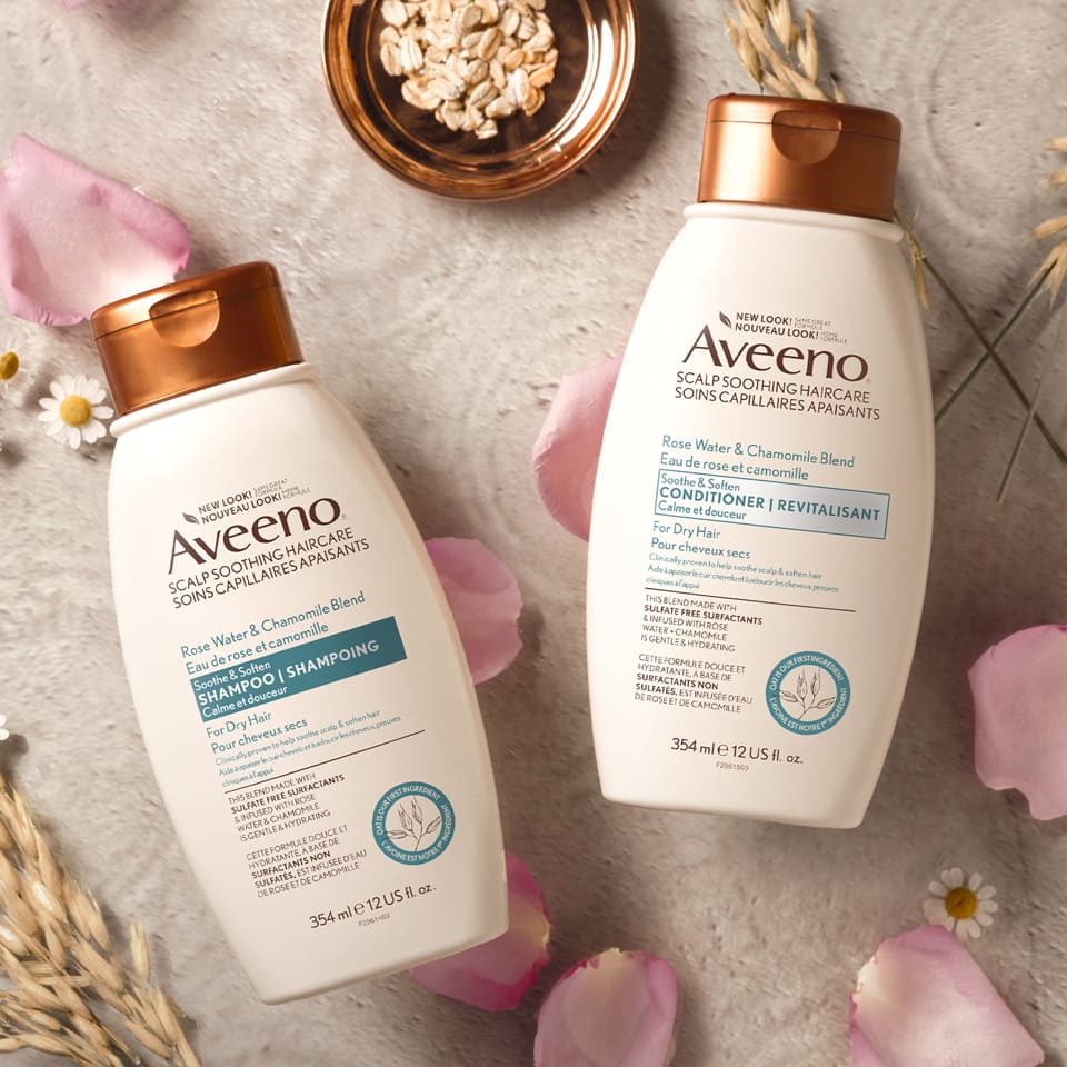 AVEENO® Rose Water and Chamomile Blend Soothe & Soften Hair Shampoo and Conditioner Bottles with oat and rose petals in the background.
